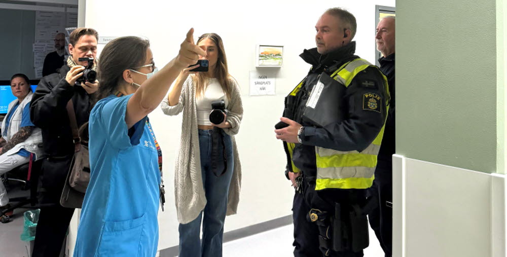 En sjuksköterska med munskydd och blå skjorta pekar bortåt med handen och pratar med en manlig polis i en korridor. Bakom dem står två personer, en ung man me dkamera och en kvinna som fotar med mobiltelefon. De är från tv och tidning. 