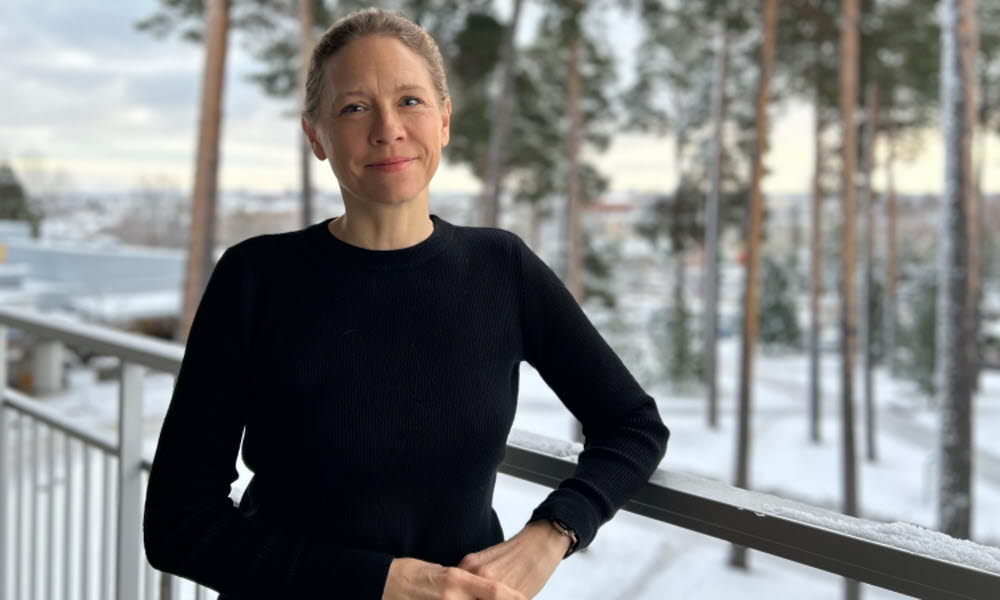 Ung kvinna står ute på en balkong, vinter ute, snö och tallar i bakgrunden. Bär svart tröja, ingen mössa och inga vantar.