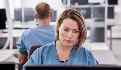 Serious female doctor using computer. Young medical professional sitting at desk in control room. She is in scrubs at hospital.