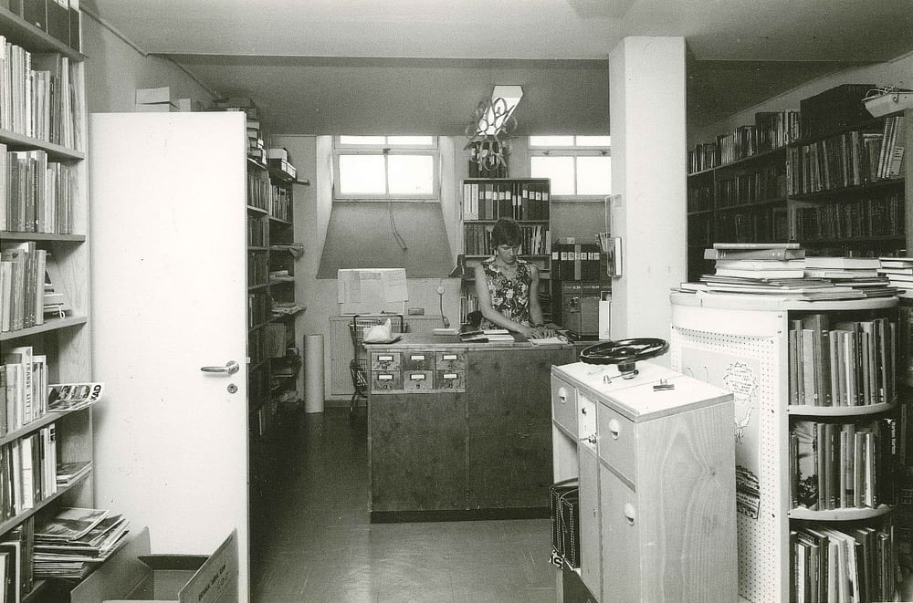 Svartvit bild av ett äldre bibliotek där en kvinna arbetar bakom en trädisk med kortkatalog. Rummet är fullt av bokhyllor, pärmar och dokument i ett trångt men organiserat utrymme.