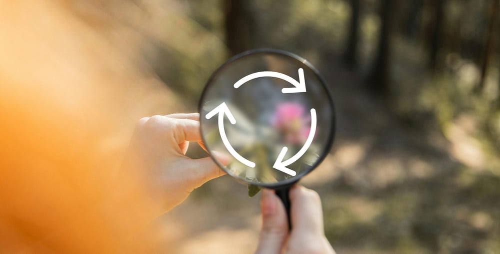 Hand som håller i ett förstoringsglas med skog i bakgrunden. I förstoringsglaset syns en illustration av cirkulär ekonomi