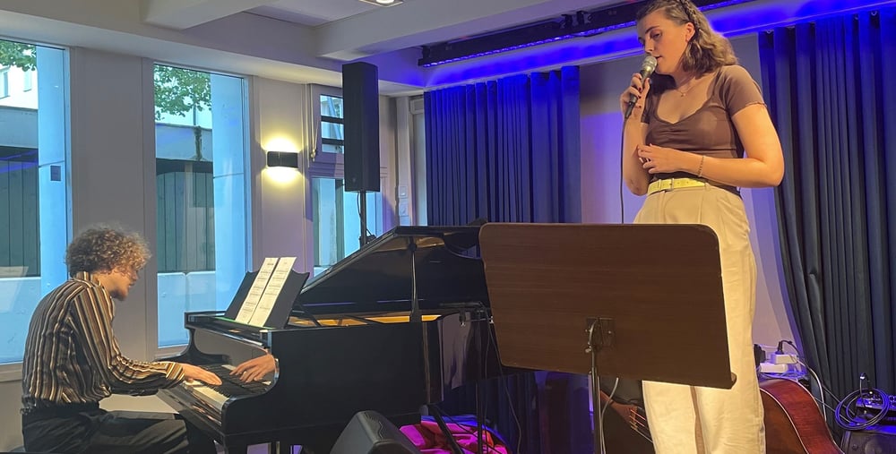 Person står på en liten scen i ett klassrum och  sjunger. En person sitter vid piano och spelar. 