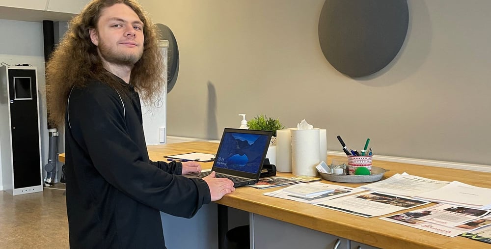 Person vid en dator ståendes vid en arbetsbänk med papper liggandes på sidan.