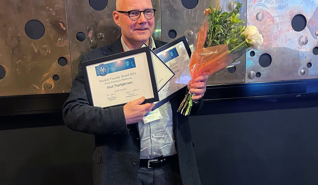 Kärlkirurgen Knut Thorbjørnsen, medelålders man står med blommor i handen och flera inramade prisdokument i handen. Han ler mot kameran. Han bär en ljus skjorta och en grå kavaj. 