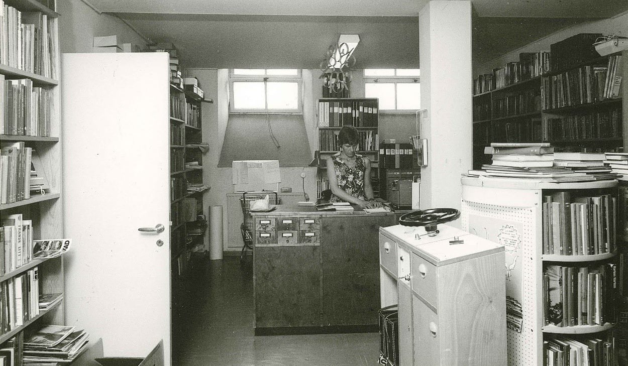 Svartvit bild av ett äldre bibliotek där en kvinna arbetar bakom en trädisk med kortkatalog. Rummet är fullt av bokhyllor, pärmar och dokument i ett trångt men organiserat utrymme.