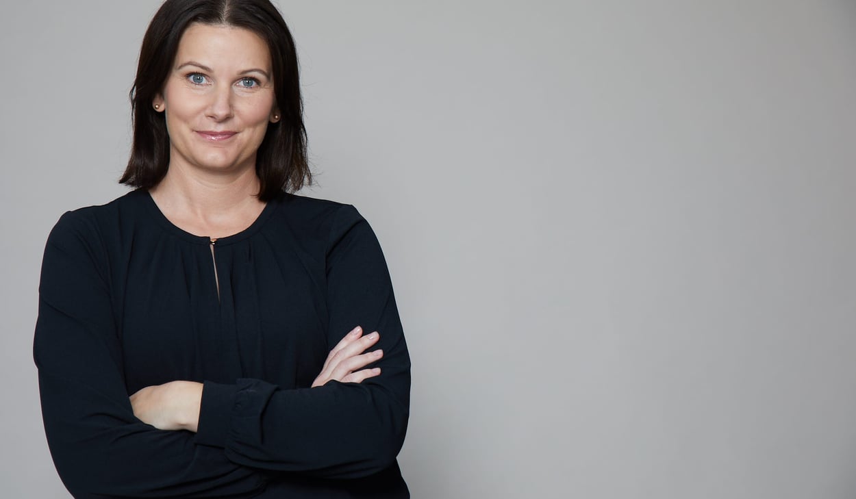 Pressbild på Anna E Wahlgren stående framför en grå vägg, klädd i svart långärmad tröja