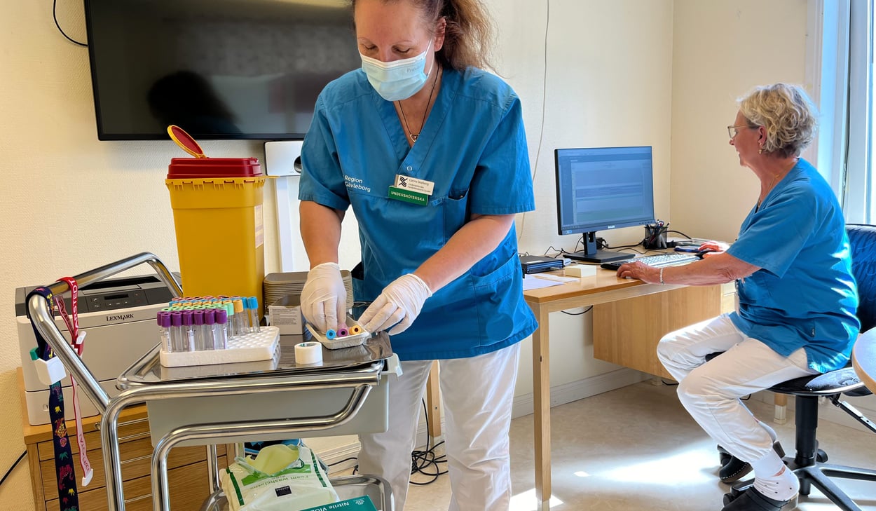 två personer i arbetskläder vid ett rullbord med provrör och vid en dator