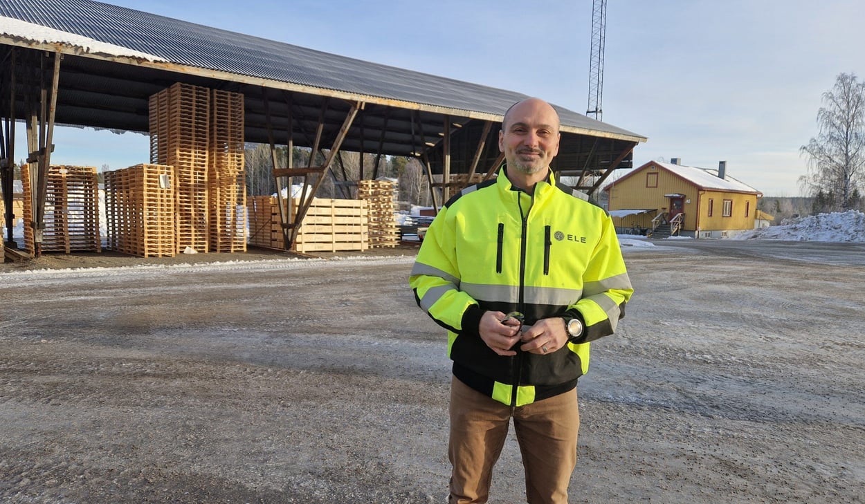 Mattias Ringström iklädd gul varseljacka står på en industriell gårdsplan med staplade träpallar och ett skjul i bakgrunden.