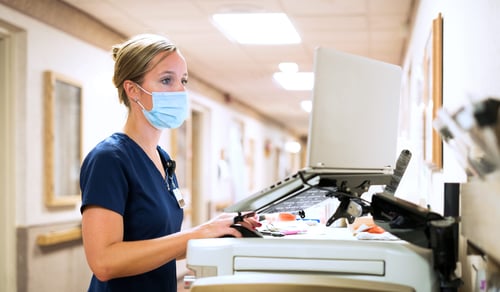 Registered nurse wearing a face mask documents medicine provided to a patient into a laptop computer at the nursing station at work, Indiana, USA