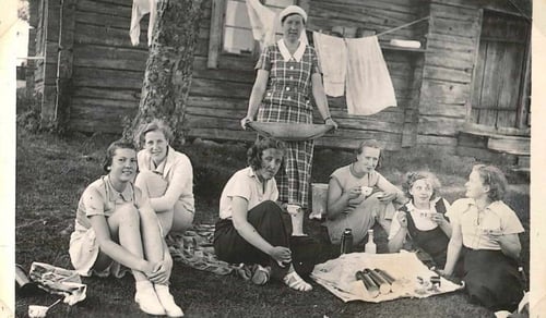 Ett svartvitt foto av en grupp människor som sitter och fikar på gräset.