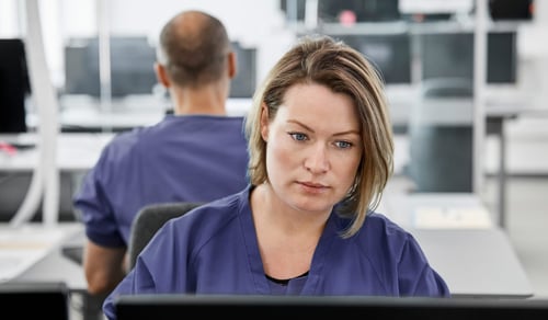 Serious female doctor using computer. Young medical professional sitting at desk in control room. She is in scrubs at hospital.