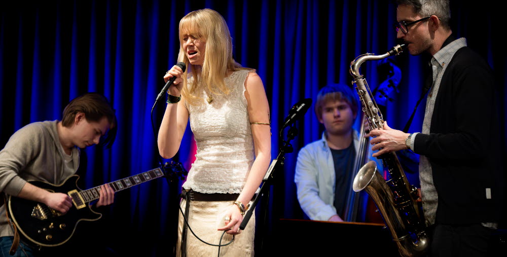 Musiker uppträder på scen med sång, gitarr, kontrabas och saxofon.