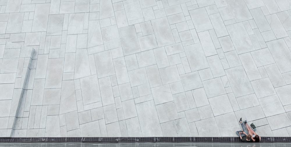 Två personer sitter på marken intill en speglande vägg på ett stort ljusgrått stenlagt torg.